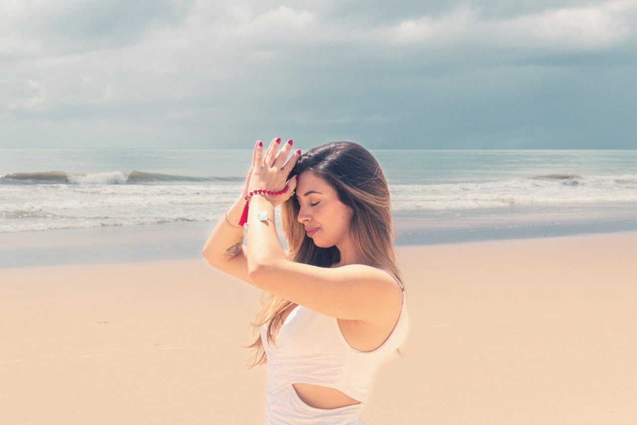 Madu na praia, de roupa clara, com as mãos unidas próximas ao rosto e o mar ao fundo