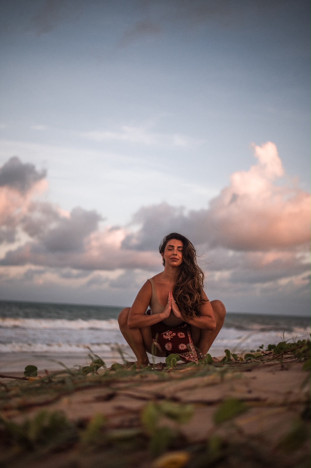 Madu agachada com as mãos em namastê, na praia ao entardecer