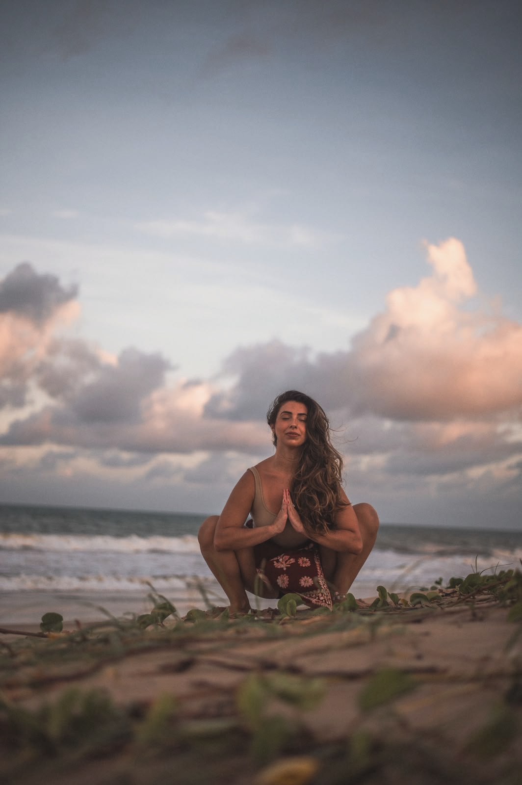 Madu agachada com as mãos em namastê, na praia ao entardecer
